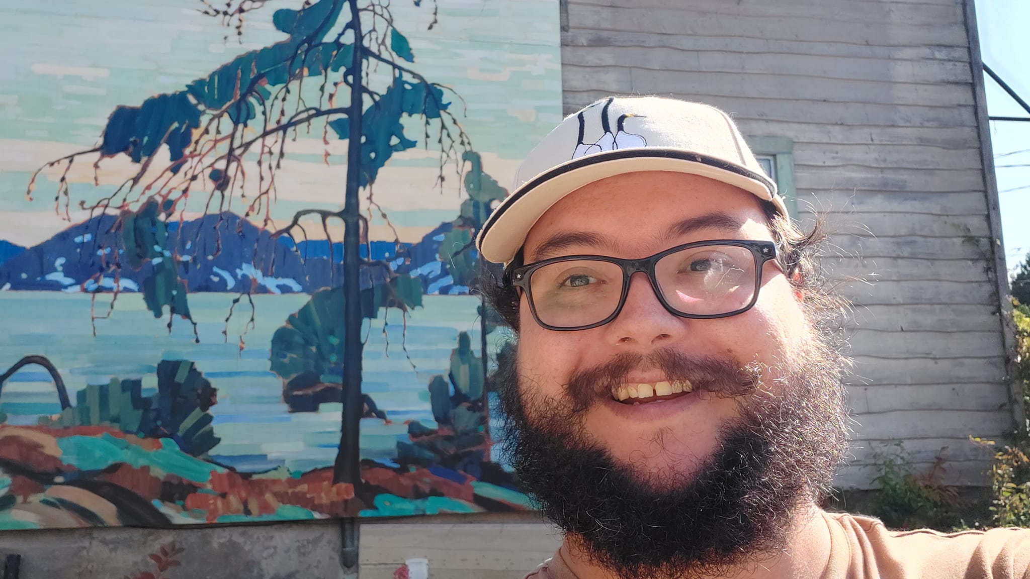 A man with short hair and bushy facial hair stands in front of an outdoor mural of Tom Thompson's painting "The Jack Pine" on the side of a wooden building, he is estatic